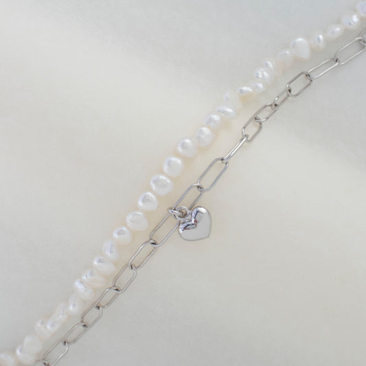 This stylish piece features a dual chain with baroque pearls and an alta chain with a heart pendant.